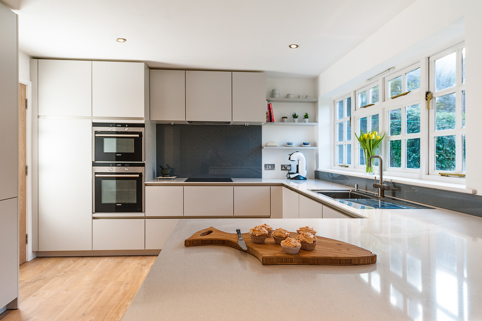 Example of a mid-sized trendy u-shaped medium tone wood floor kitchen design in Hampshire with an integrated sink, flat-panel cabinets, gray cabinets, solid surface countertops, white backsplash, glass sheet backsplash, stainless steel appliances and a peninsula