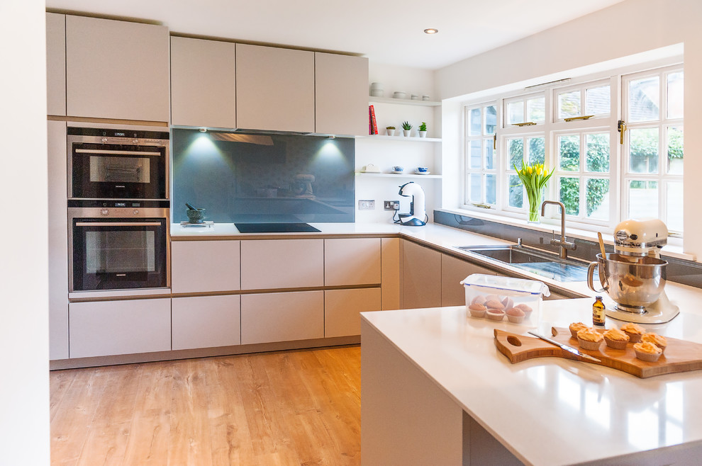 Medium sized contemporary u-shaped kitchen in Hampshire with an integrated sink, flat-panel cabinets, grey cabinets, composite countertops, white splashback, glass sheet splashback, stainless steel appliances, medium hardwood flooring and a breakfast bar.