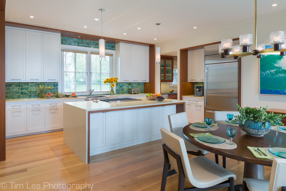 Design ideas for a traditional l-shaped kitchen/diner in Bridgeport with a belfast sink, shaker cabinets, white cabinets, engineered stone countertops, green splashback, glass tiled splashback, stainless steel appliances, light hardwood flooring, an island and brown floors.