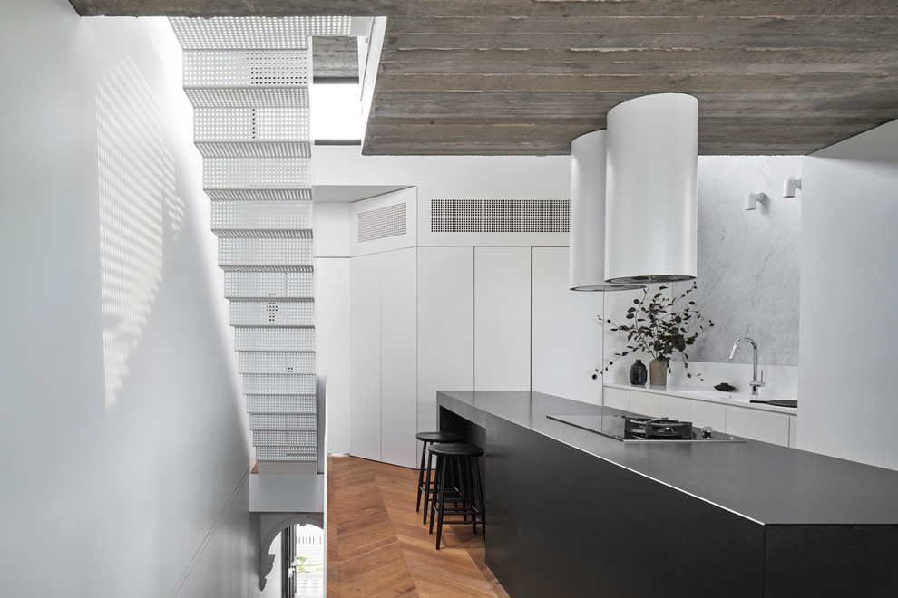 Small contemporary galley open plan kitchen in Melbourne with a submerged sink, flat-panel cabinets, white cabinets, granite worktops, grey splashback, marble splashback, black appliances, medium hardwood flooring, an island, brown floors and black worktops.