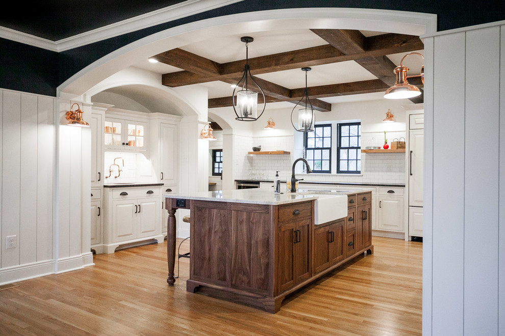 Example of a classic u-shaped medium tone wood floor and brown floor enclosed kitchen design in Other with a farmhouse sink, raised-panel cabinets, white cabinets, white backsplash, subway tile backsplash, paneled appliances, an island and white countertops
