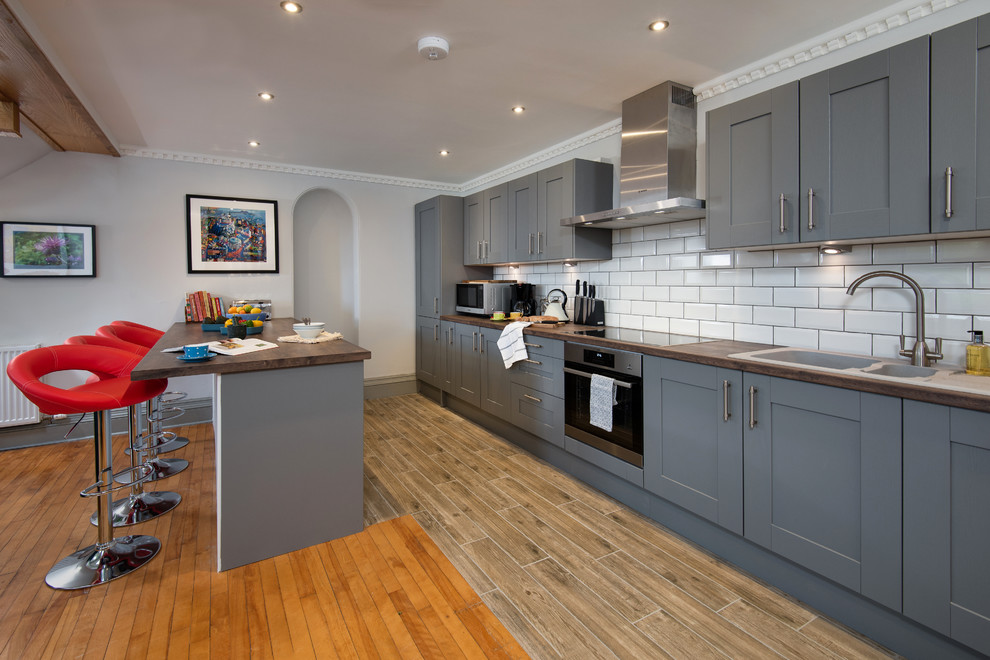 Photo of a classic galley kitchen in Other with shaker cabinets, grey cabinets, wood worktops, white splashback, metro tiled splashback, stainless steel appliances, a breakfast bar, beige floors and brown worktops.