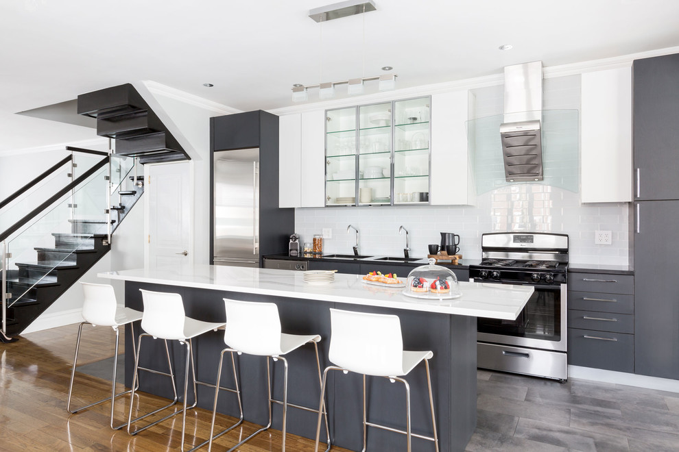 Example of a trendy gray floor open concept kitchen design in New York with an island, a double-bowl sink, glass-front cabinets, stainless steel cabinets, white backsplash, subway tile backsplash, stainless steel appliances and black countertops