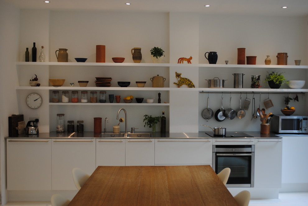 This is an example of a small contemporary single-wall kitchen/diner in London with a double-bowl sink, flat-panel cabinets, white cabinets, stainless steel worktops, white splashback, glass sheet splashback, stainless steel appliances, concrete flooring and no island.