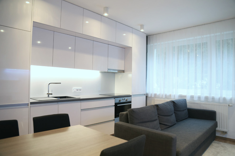 Photo of a small modern single-wall open plan kitchen in Other with a built-in sink, flat-panel cabinets, white cabinets, laminate countertops, white splashback, glass sheet splashback, stainless steel appliances, light hardwood flooring, no island and brown floors.