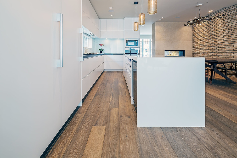 Example of a small trendy galley medium tone wood floor kitchen design in Toronto with an integrated sink, flat-panel cabinets, white cabinets, white backsplash, glass sheet backsplash, stainless steel appliances and an island