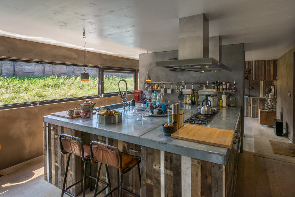 Bild på ett litet industriellt kök, med en integrerad diskho, släta luckor, skåp i slitet trä, bänkskiva i rostfritt stål, mellanmörkt trägolv och en köksö