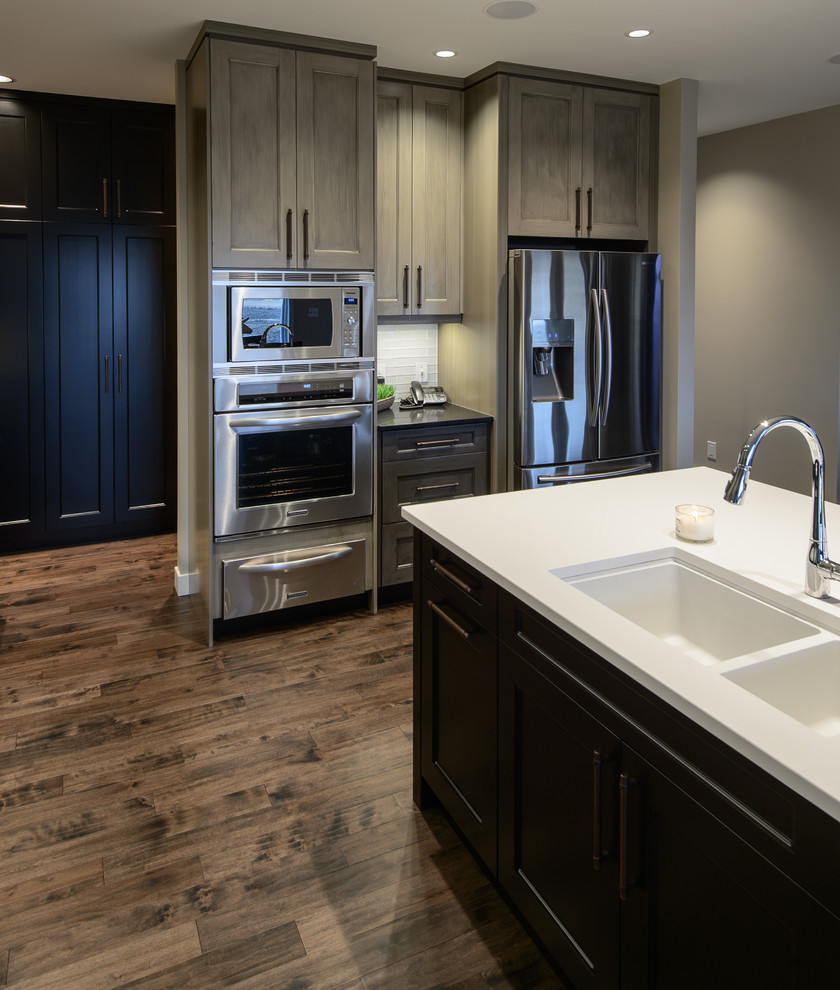 Example of a mid-sized transitional l-shaped dark wood floor and brown floor open concept kitchen design in Other with a double-bowl sink, shaker cabinets, gray cabinets, solid surface countertops, white backsplash, porcelain backsplash, stainless steel appliances and an island
