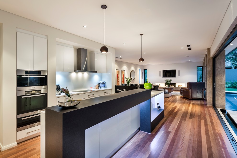 Eat-in kitchen - mid-sized contemporary galley medium tone wood floor and brown floor eat-in kitchen idea in Perth with a double-bowl sink, recessed-panel cabinets, white cabinets, quartz countertops, white backsplash, glass sheet backsplash, stainless steel appliances, an island and white countertops