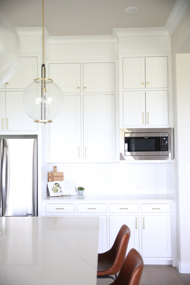 Foto på ett mellanstort vintage vit linjärt kök och matrum, med en undermonterad diskho, skåp i shakerstil, vita skåp, bänkskiva i kvarts, vitt stänkskydd, stänkskydd i keramik, rostfria vitvaror, ljust trägolv, en köksö och brunt golv