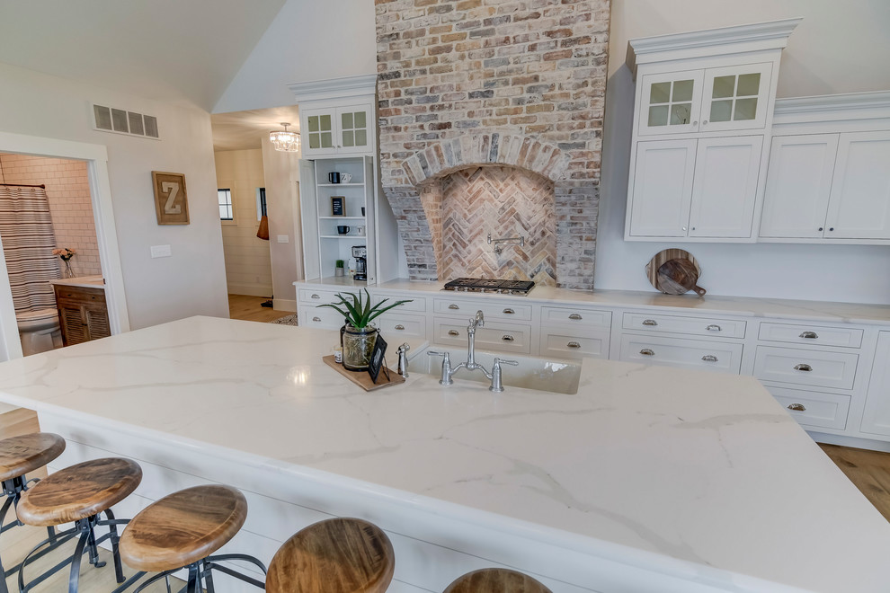 Photo of a large farmhouse l-shaped open plan kitchen in Chicago with a belfast sink, flat-panel cabinets, white cabinets, quartz worktops, stainless steel appliances, light hardwood flooring, an island and white worktops.