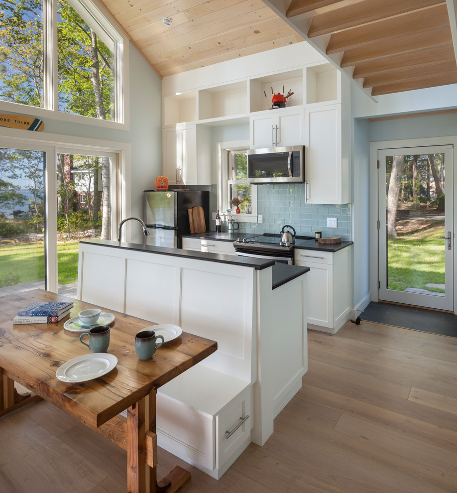 Aménagement d'une petite cuisine ouverte bord de mer avec un évier encastré, un placard à porte shaker, des portes de placard blanches, un plan de travail en granite, une crédence bleue, une crédence en carreau de verre, un électroménager en acier inoxydable, un sol en bois brun, îlot et un sol marron.