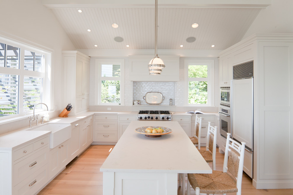 Klassische Küche in U-Form mit Landhausspüle, Schrankfronten im Shaker-Stil, weißen Schränken, Küchenrückwand in Weiß, Elektrogeräten mit Frontblende, hellem Holzboden und Kücheninsel in Vancouver