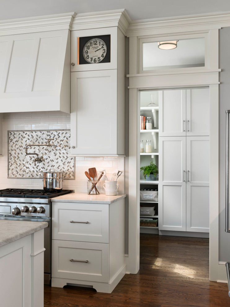 This is an example of a large traditional u-shaped kitchen pantry with a submerged sink, shaker cabinets, white cabinets, quartz worktops, white splashback, metro tiled splashback, stainless steel appliances, medium hardwood flooring, an island, brown floors and white worktops.