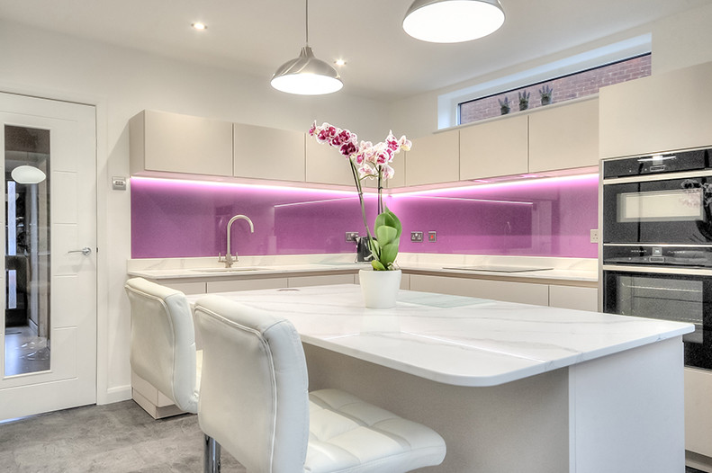 Modern inredning av ett kök, med en enkel diskho, släta luckor, vita skåp, bänkskiva i kvartsit, glaspanel som stänkskydd, svarta vitvaror, vinylgolv, en köksö, grått golv och rosa stänkskydd