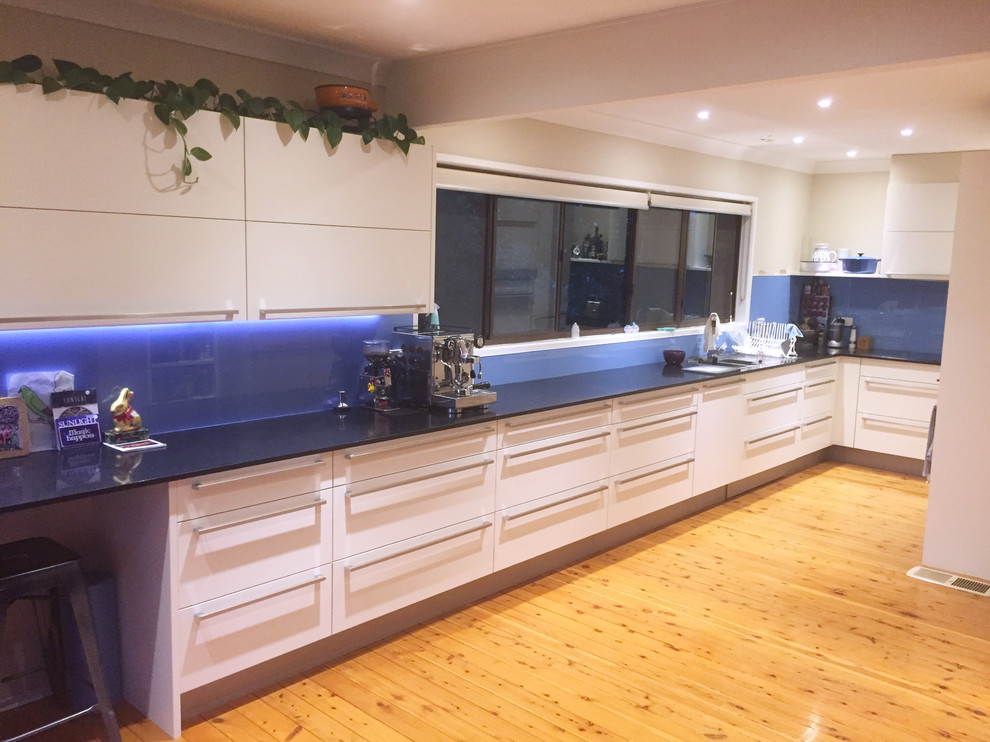 Immagine di una grande cucina a L design con ante lisce, ante bianche, paraspruzzi blu, paraspruzzi con lastra di vetro e top nero