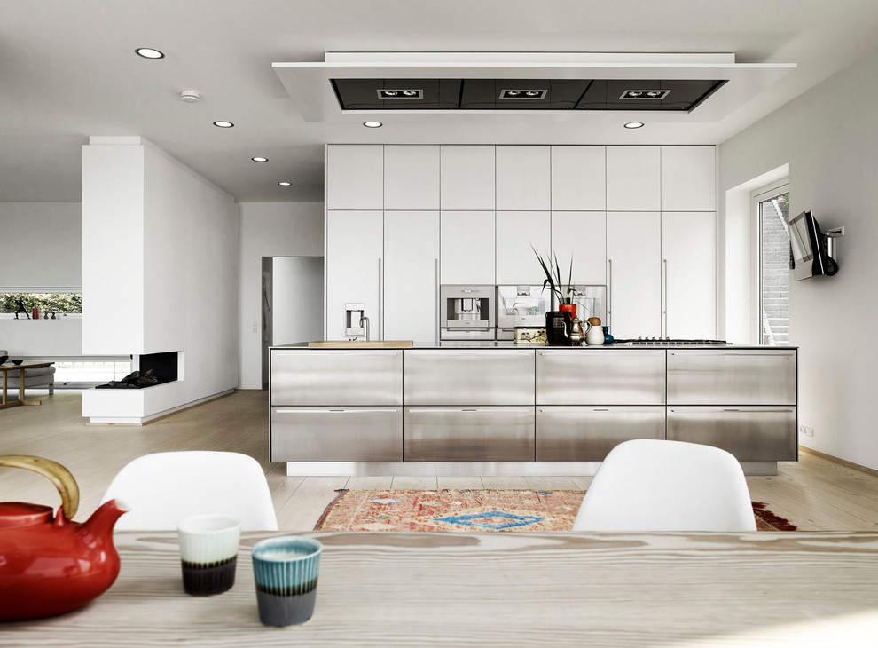 This is an example of a large contemporary galley open plan kitchen in London with flat-panel cabinets, white cabinets, stainless steel worktops, stainless steel appliances, light hardwood flooring and an island.