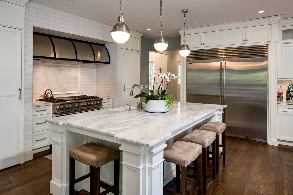 This is an example of a classic l-shaped kitchen in Detroit with a belfast sink, recessed-panel cabinets, white cabinets, white splashback, metro tiled splashback, stainless steel appliances, dark hardwood flooring and an island.