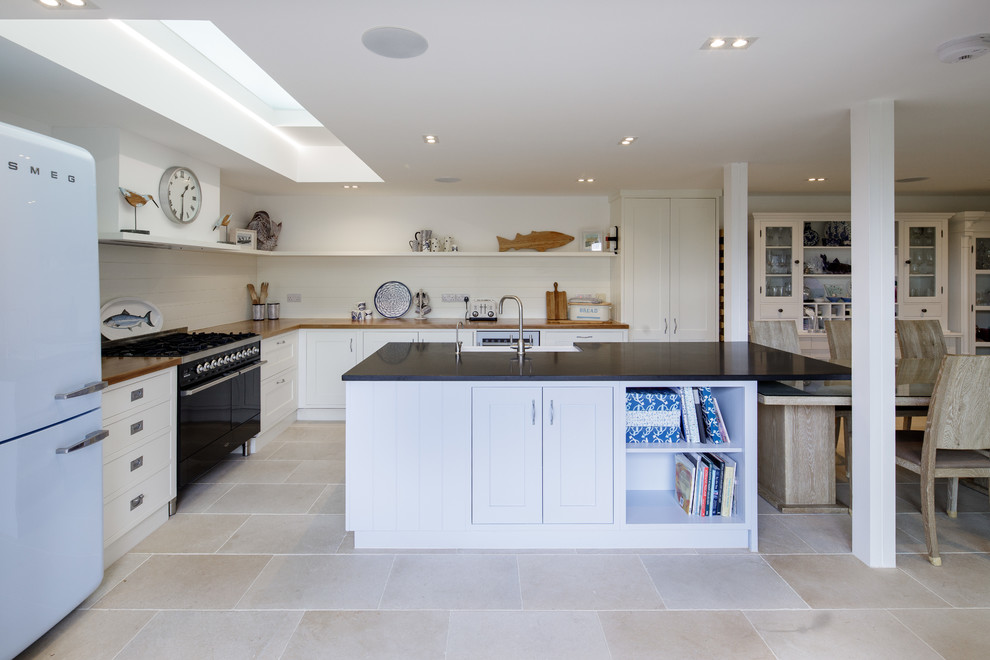 This is an example of a large coastal l-shaped kitchen in Channel Islands with a belfast sink, recessed-panel cabinets, black appliances, an island, beige floors, black worktops and white cabinets.
