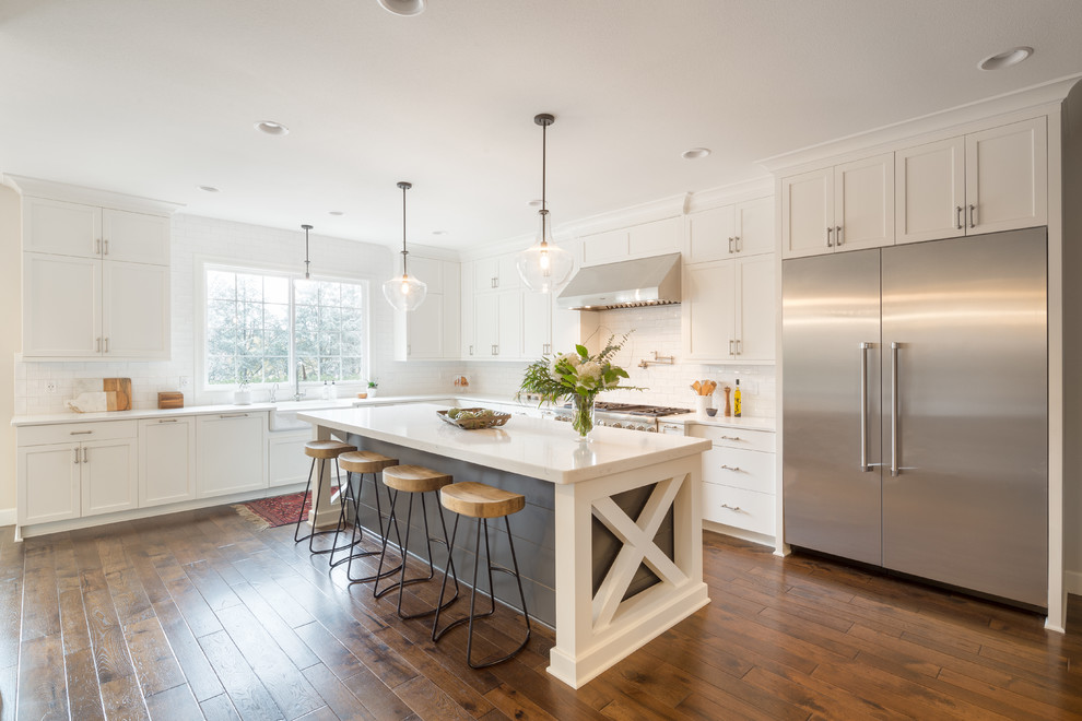 This is an example of a classic l-shaped kitchen in Portland with a belfast sink, shaker cabinets, white cabinets, white splashback, metro tiled splashback, stainless steel appliances, an island and dark hardwood flooring.