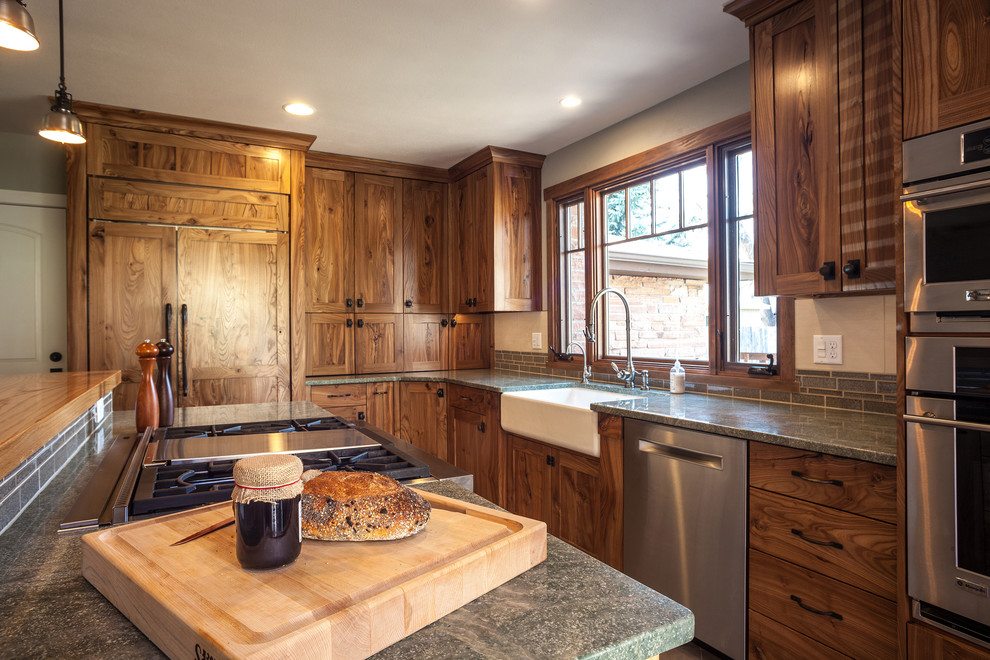 Amerikansk inredning av ett mellanstort kök, med en rustik diskho, luckor med infälld panel, skåp i mellenmörkt trä, granitbänkskiva, grönt stänkskydd, stänkskydd i glaskakel, integrerade vitvaror, klinkergolv i porslin och en köksö