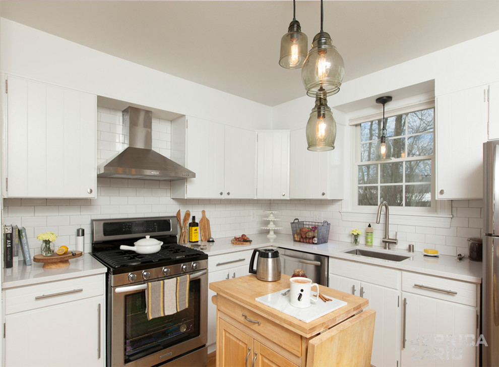 Example of a small cottage u-shaped medium tone wood floor and brown floor enclosed kitchen design with a drop-in sink, flat-panel cabinets, white cabinets, quartz countertops, white backsplash, ceramic backsplash, stainless steel appliances and an island