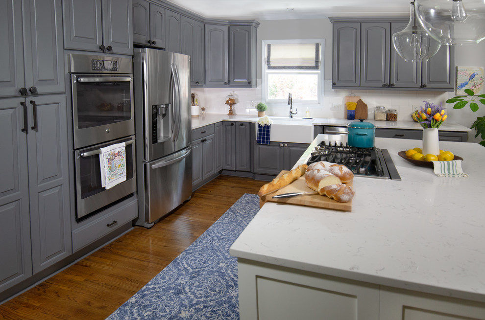 Rustik inredning av ett stort vit vitt kök, med en rustik diskho, luckor med upphöjd panel, grå skåp, bänkskiva i kvarts, vitt stänkskydd, stänkskydd i keramik, rostfria vitvaror, mellanmörkt trägolv, en köksö och brunt golv