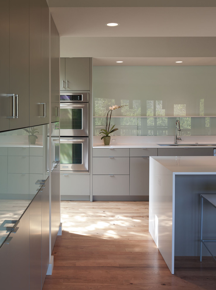 Example of a mid-sized minimalist galley medium tone wood floor and brown floor eat-in kitchen design in Austin with stainless steel appliances, flat-panel cabinets, gray cabinets, quartz countertops, glass sheet backsplash, an undermount sink and an island