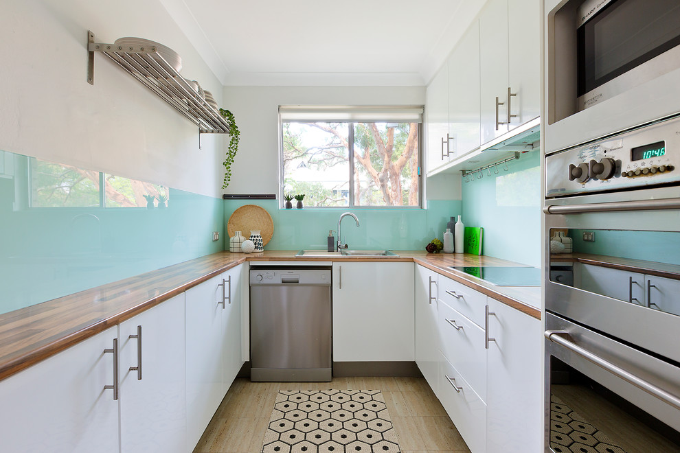 シドニーにある小さなコンテンポラリースタイルのおしゃれなキッチン (白いキャビネット、木材カウンター、青いキッチンパネル、ガラス板のキッチンパネル、シルバーの調理設備、ラミネートの床、ベージュの床、フラットパネル扉のキャビネット) の写真