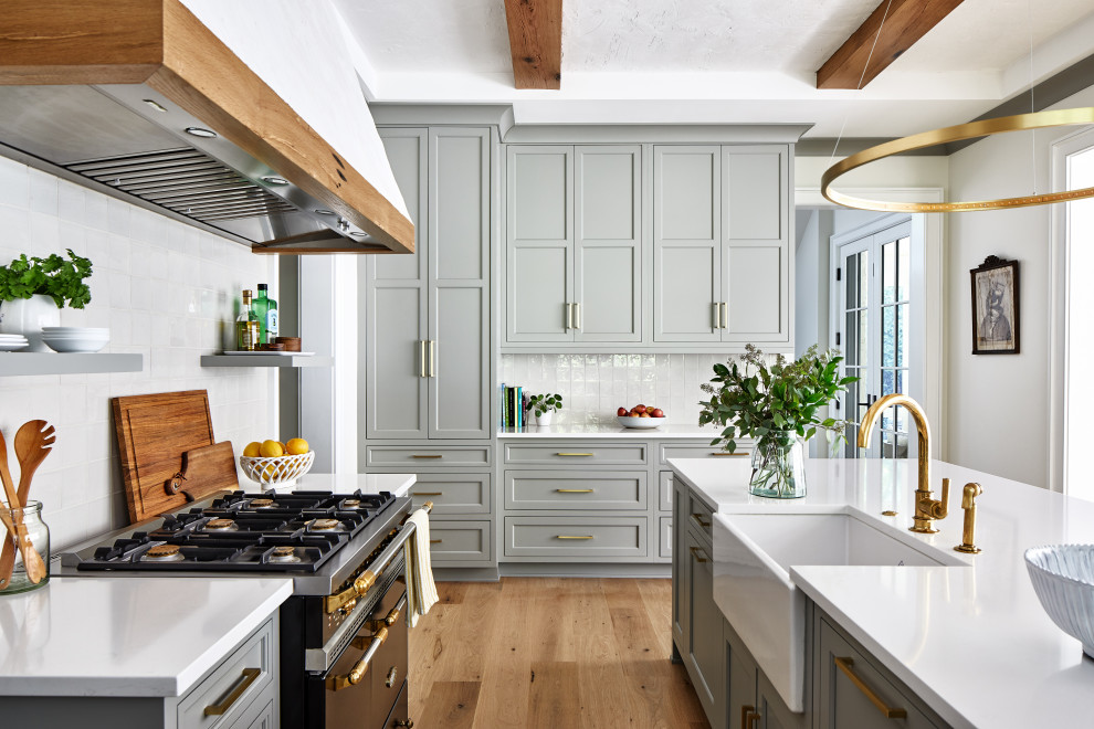 Inredning av ett klassiskt stort vit vitt l-kök, med en rustik diskho, skåp i shakerstil, grå skåp, bänkskiva i kvarts, vitt stänkskydd, stänkskydd i stenkakel, svarta vitvaror, en köksö, mellanmörkt trägolv och brunt golv