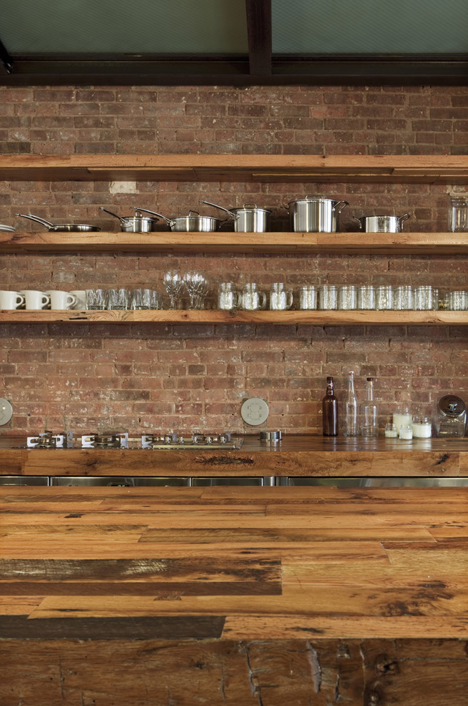 Mid-sized urban l-shaped concrete floor eat-in kitchen photo in New York with wood countertops, a farmhouse sink, flat-panel cabinets, stainless steel cabinets, red backsplash, brick backsplash, stainless steel appliances and an island
