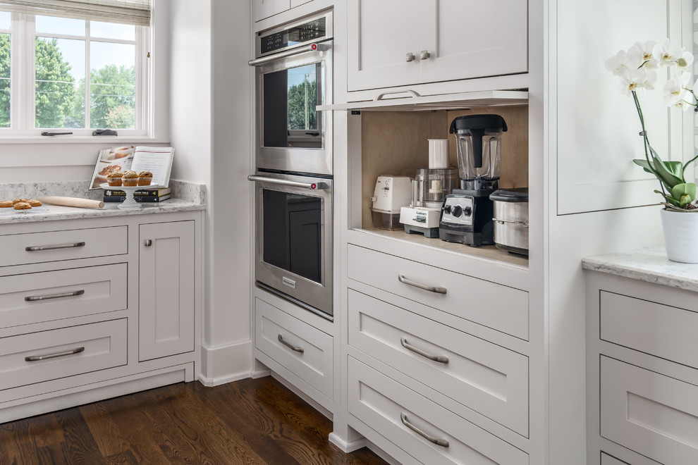 Foto på ett stort lantligt vit l-kök, med en enkel diskho, skåp i shakerstil, vita skåp, bänkskiva i kvarts, vitt stänkskydd, stänkskydd i porslinskakel, integrerade vitvaror, mörkt trägolv och en köksö