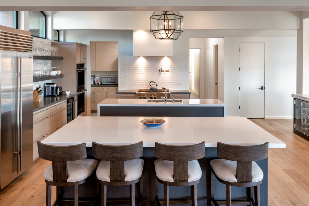 Example of a trendy l-shaped medium tone wood floor and brown floor open concept kitchen design in San Francisco with an undermount sink, flat-panel cabinets, medium tone wood cabinets, white backsplash, subway tile backsplash, stainless steel appliances, two islands and white countertops