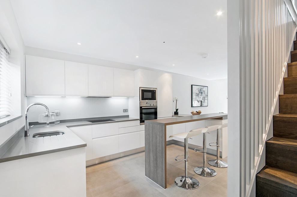 Photo of an expansive contemporary u-shaped open plan kitchen in London with flat-panel cabinets, white cabinets, quartz worktops, white splashback, glass sheet splashback, stainless steel appliances and porcelain flooring.