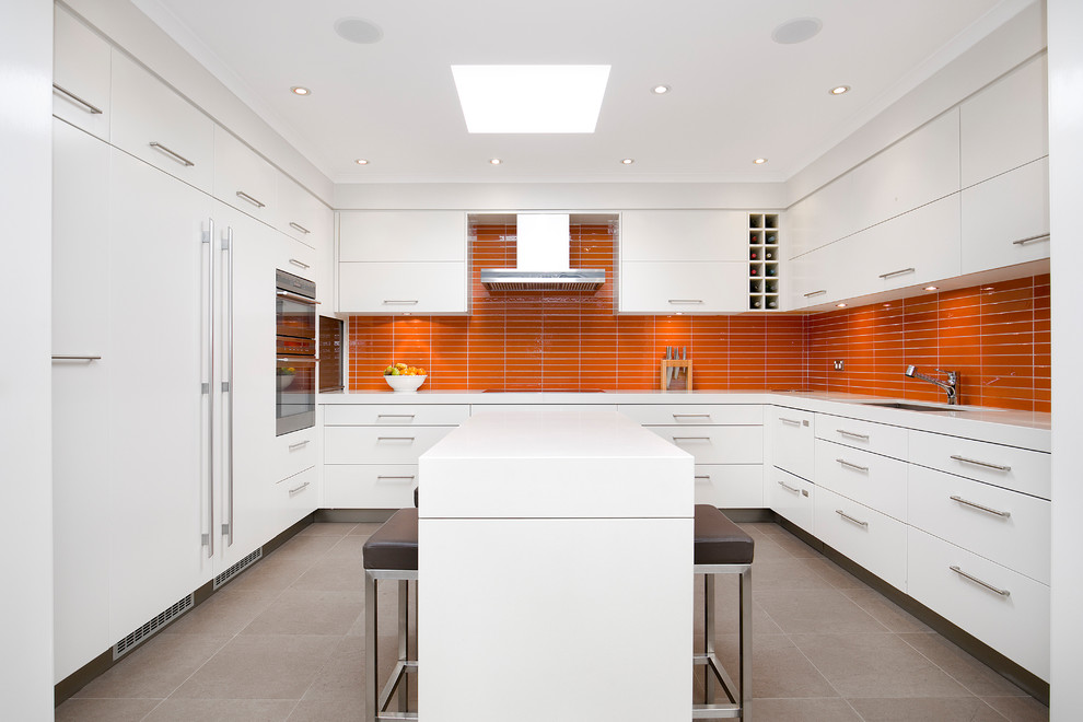 На фото: п-образная кухня в современном стиле с двойной мойкой, оранжевым фартуком, техникой под мебельный фасад, полом из керамогранита и островом с