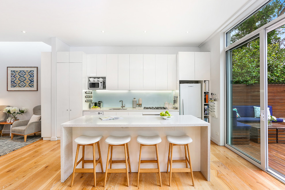 Bild på ett vintage vit linjärt vitt kök, med en undermonterad diskho, släta luckor, vita skåp, granitbänkskiva, grönt stänkskydd, glaspanel som stänkskydd, rostfria vitvaror, mellanmörkt trägolv, en köksö och brunt golv