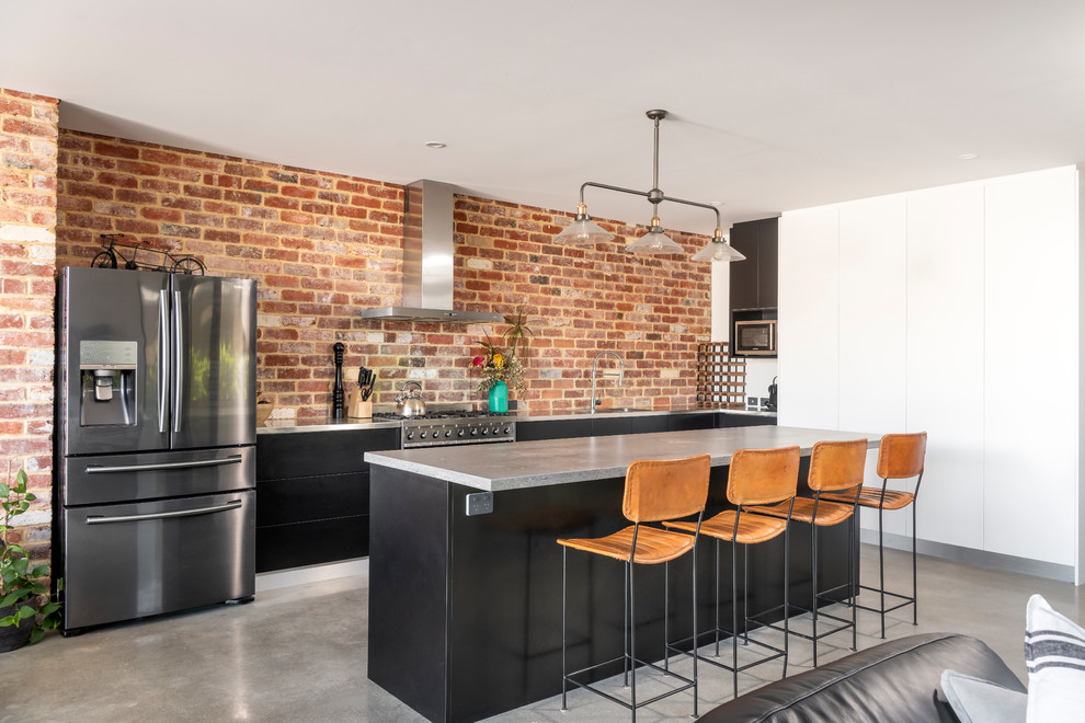 This is an example of a l-shaped open plan kitchen in Perth with flat-panel cabinets, black cabinets, brown splashback, brick splashback, stainless steel appliances, concrete flooring, an island, grey floors and grey worktops.