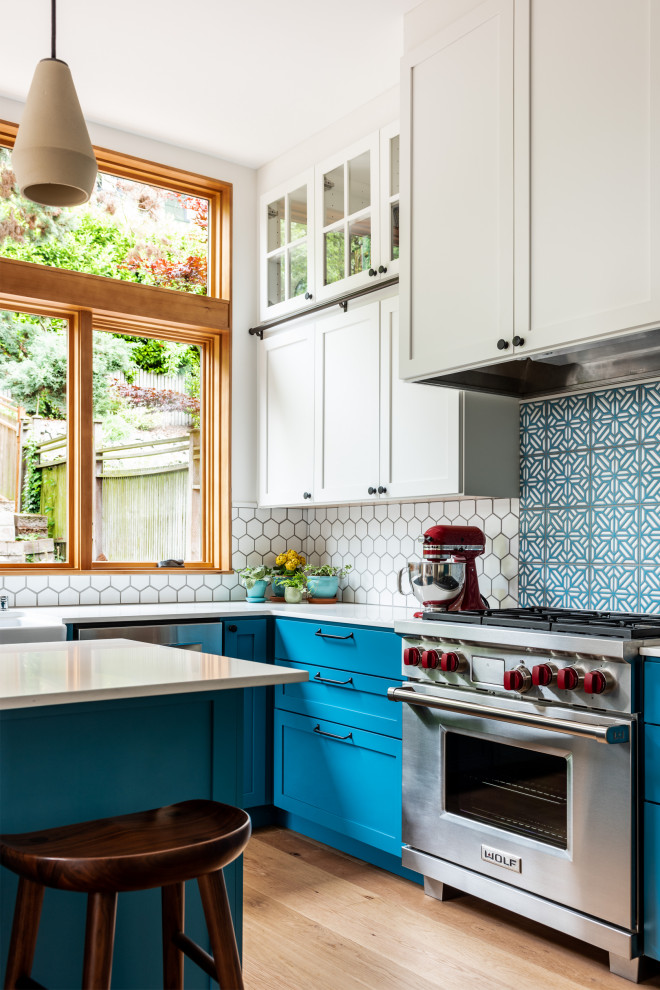 Foto de cocina actual grande con fregadero sobremueble, armarios estilo shaker, puertas de armario azules, encimera de cuarzo compacto, salpicadero blanco, salpicadero de azulejos de cerámica, electrodomésticos de acero inoxidable, suelo de madera clara, una isla y encimeras blancas