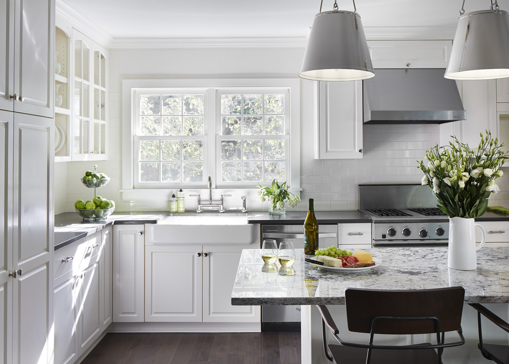 Ejemplo de cocina tradicional, de tamaño medio, con fregadero sobremueble, puertas de armario blancas, suelo de madera oscura, una isla, armarios con paneles con relieve, encimera de granito, salpicadero blanco, salpicadero de azulejos de cerámica, electrodomésticos de acero inoxidable y suelo marrón