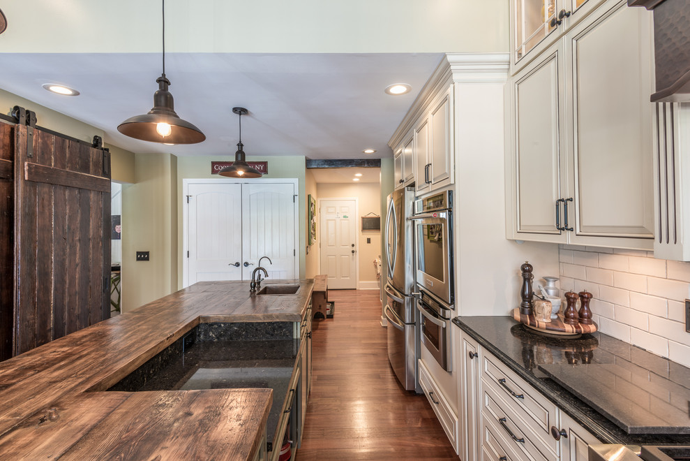 Example of a large country u-shaped medium tone wood floor and brown floor enclosed kitchen design in Other with a farmhouse sink, raised-panel cabinets, beige cabinets, quartz countertops, white backsplash, ceramic backsplash, stainless steel appliances, an island and brown countertops