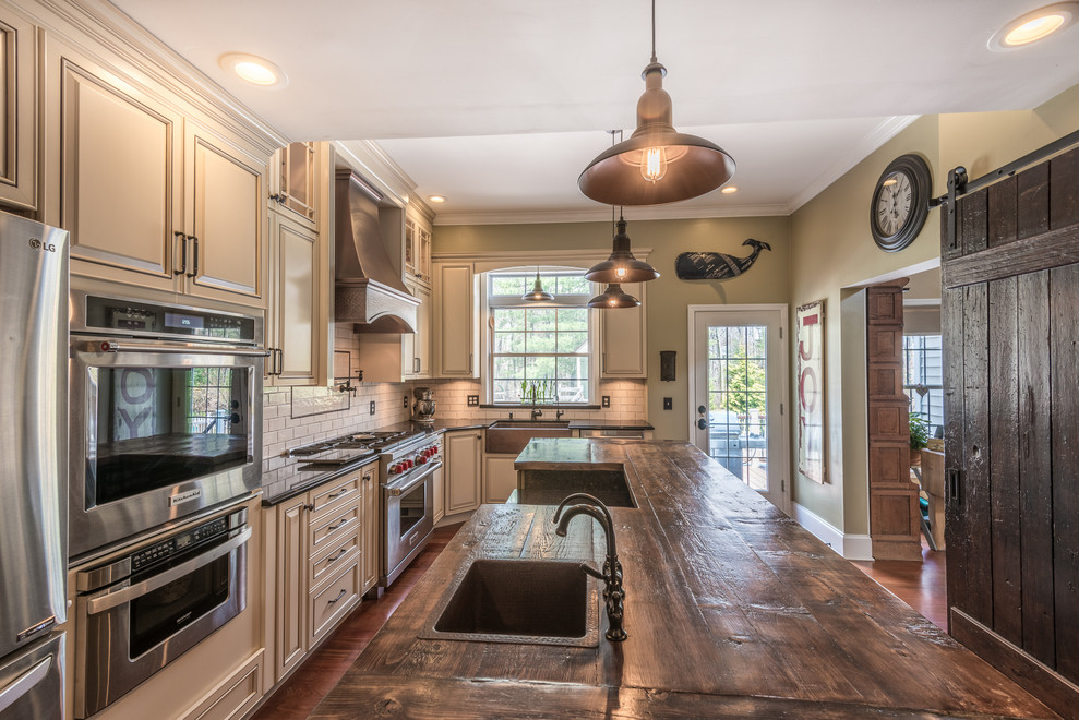 Example of a large farmhouse u-shaped medium tone wood floor and brown floor enclosed kitchen design in Other with a farmhouse sink, raised-panel cabinets, beige cabinets, quartz countertops, white backsplash, ceramic backsplash, stainless steel appliances, an island and brown countertops