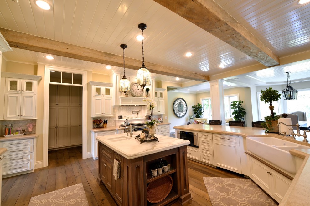 Mid-sized elegant u-shaped medium tone wood floor and brown floor open concept kitchen photo in Other with a farmhouse sink, recessed-panel cabinets, white cabinets, marble countertops, gray backsplash, stainless steel appliances and an island