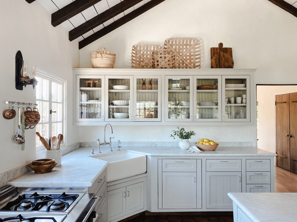 Lantlig inredning av ett vit vitt l-kök, med en rustik diskho, skåp i shakerstil, vita skåp, mörkt trägolv, en köksö och brunt golv