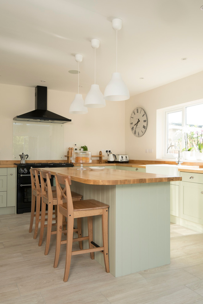 This is an example of a large traditional l-shaped kitchen/diner in Cardiff with a belfast sink, shaker cabinets, green cabinets, wood worktops, glass sheet splashback, black appliances, ceramic flooring and an island.