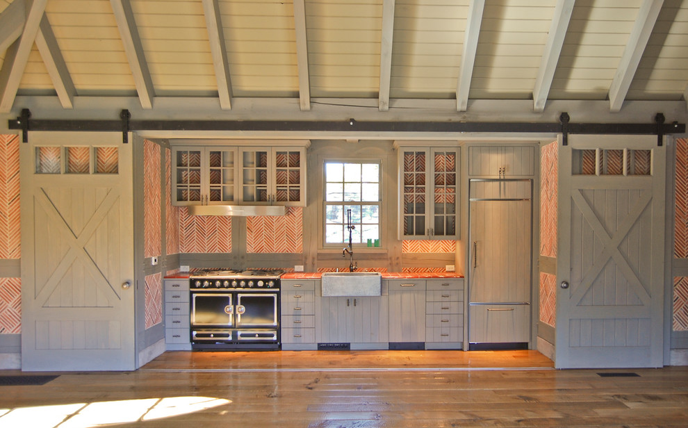 Bild på ett litet lantligt linjärt kök med öppen planlösning, med en rustik diskho, släta luckor, grå skåp, orange stänkskydd, stänkskydd i glaskakel, rostfria vitvaror, mellanmörkt trägolv och brunt golv