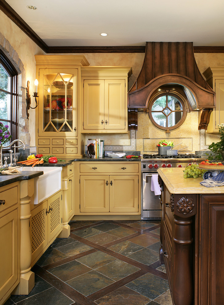 Bild på ett stort kök, med luckor med profilerade fronter, en rustik diskho, gula skåp, bänkskiva i täljsten, gult stänkskydd, rostfria vitvaror, skiffergolv, stänkskydd i mosaik, en köksö och brunt golv