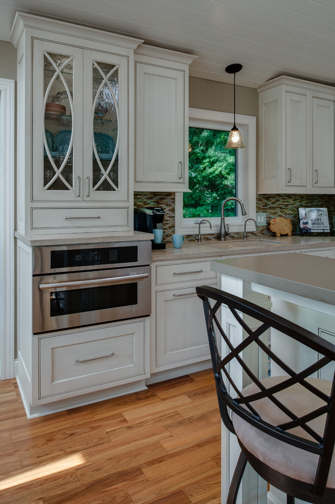 Example of a mid-sized classic l-shaped medium tone wood floor eat-in kitchen design in Other with an integrated sink, recessed-panel cabinets, gray cabinets, solid surface countertops, multicolored backsplash, glass sheet backsplash, stainless steel appliances and an island