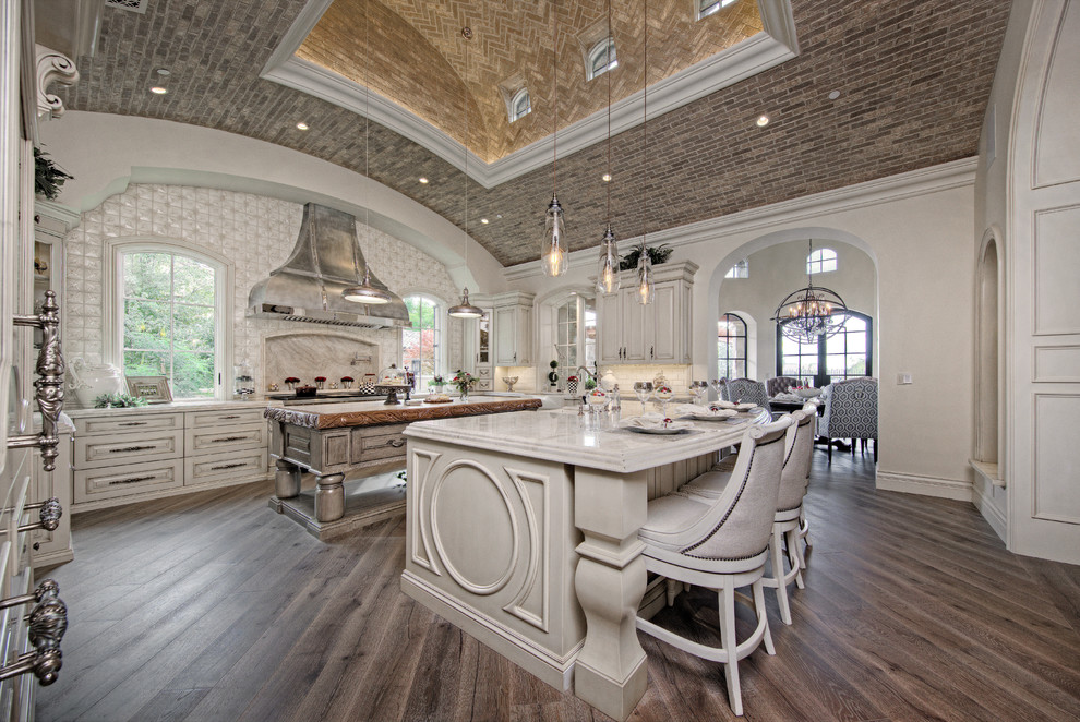 Huge transitional u-shaped medium tone wood floor enclosed kitchen photo in Phoenix with a farmhouse sink, raised-panel cabinets, white cabinets, marble countertops, white backsplash, mosaic tile backsplash, stainless steel appliances and two islands