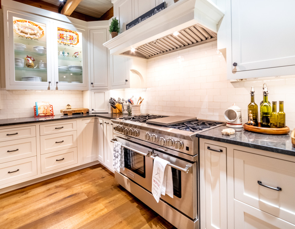 Diseño de cocina comedor de estilo americano con puertas de armario blancas, salpicadero blanco, electrodomésticos de acero inoxidable y suelo marrón