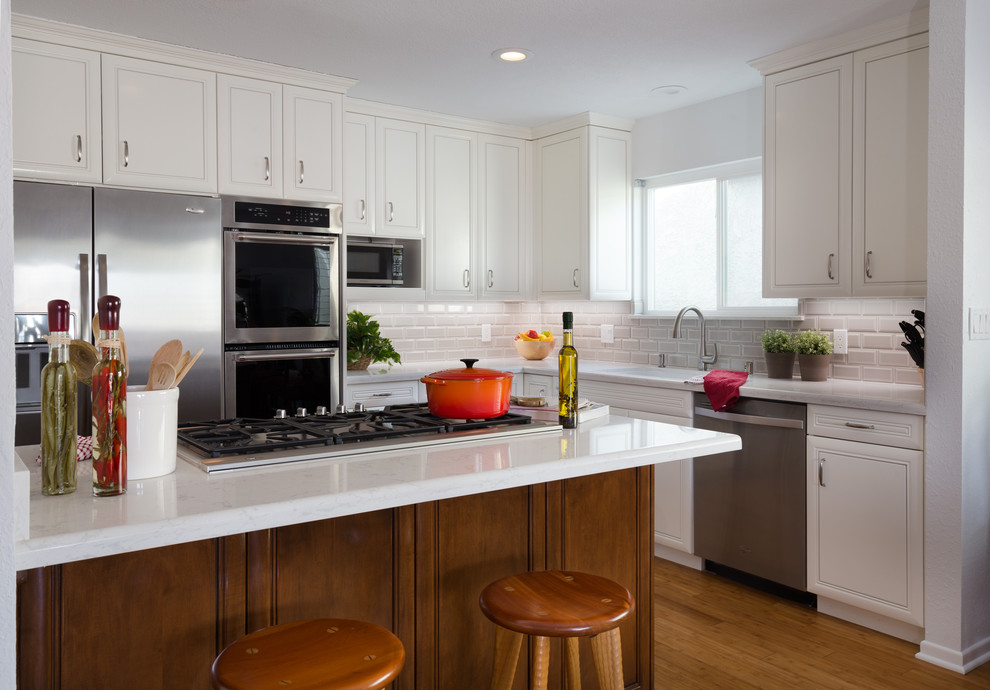 This is an example of a medium sized classic l-shaped kitchen/diner in Los Angeles with white cabinets, engineered stone countertops, white splashback, stainless steel appliances, medium hardwood flooring, a breakfast bar, a submerged sink, beaded cabinets, metro tiled splashback and brown floors.